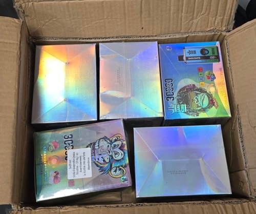 A cardboard box containing shiny holographic packaging, including a visible box labeled "Strawberry Kiwi" and "Blueberry Raspberry Lemonade" with colorful graphics and illustrations.